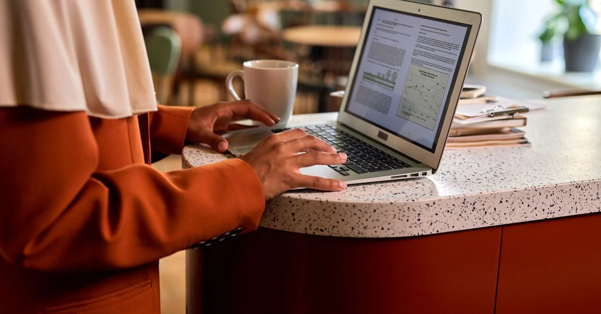 Femme adulte tapant sur un ordinateur portable à un comptoir de bureau moderne, avec un café à côté d'elle dans un cadre intérieur décontracté.