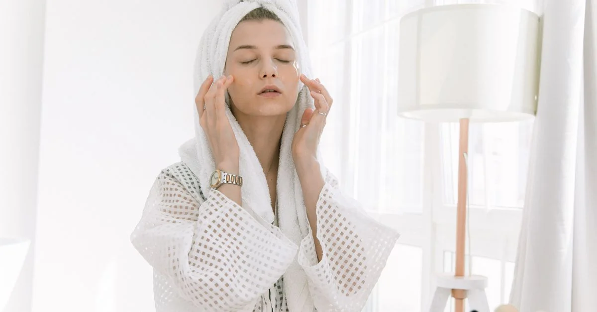 Femme adulte en peignoir effectuant une routine de soins de la peau avec une serviette sur la tête et des patchs pour les yeux à l'intérieur.