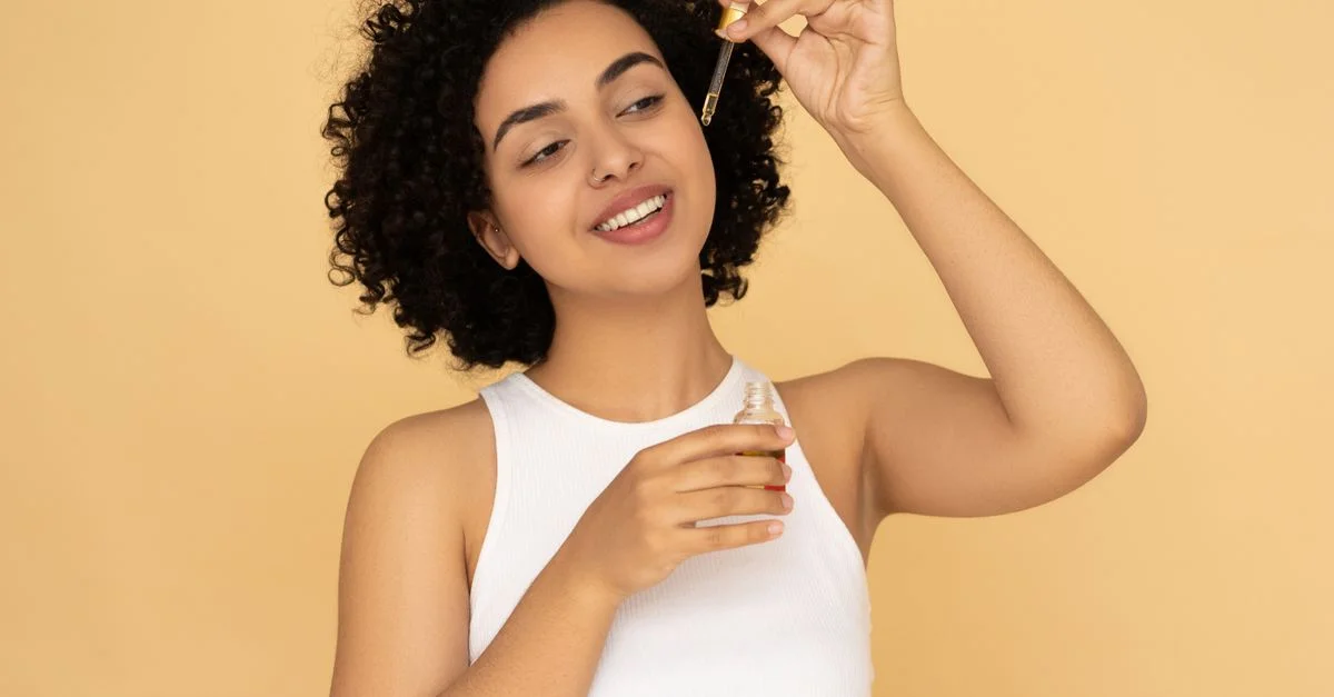 Une femme aux cheveux bouclés applique de l'huile essentielle sur sa peau, favorisant le bien-être et le soin de la peau.