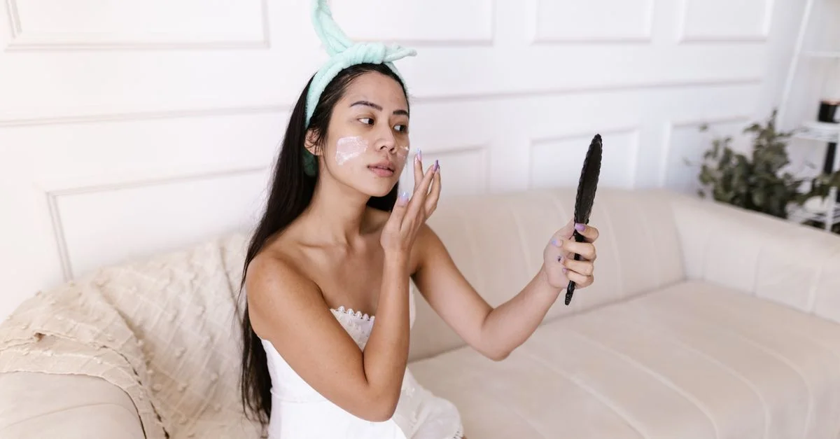 Femme asiatique en serviette appliquant de la crème pour le visage tout en se regardant dans un miroir de poche à l'intérieur.